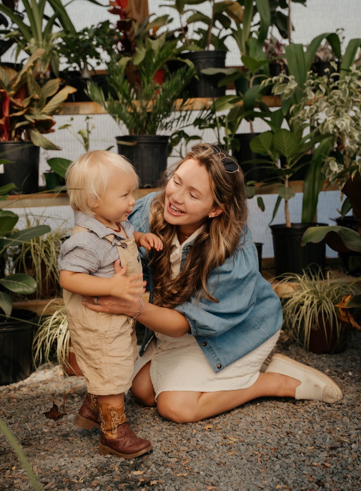 Mother kneeling on gravel beside her toddler in a greenhouse full of potted plants