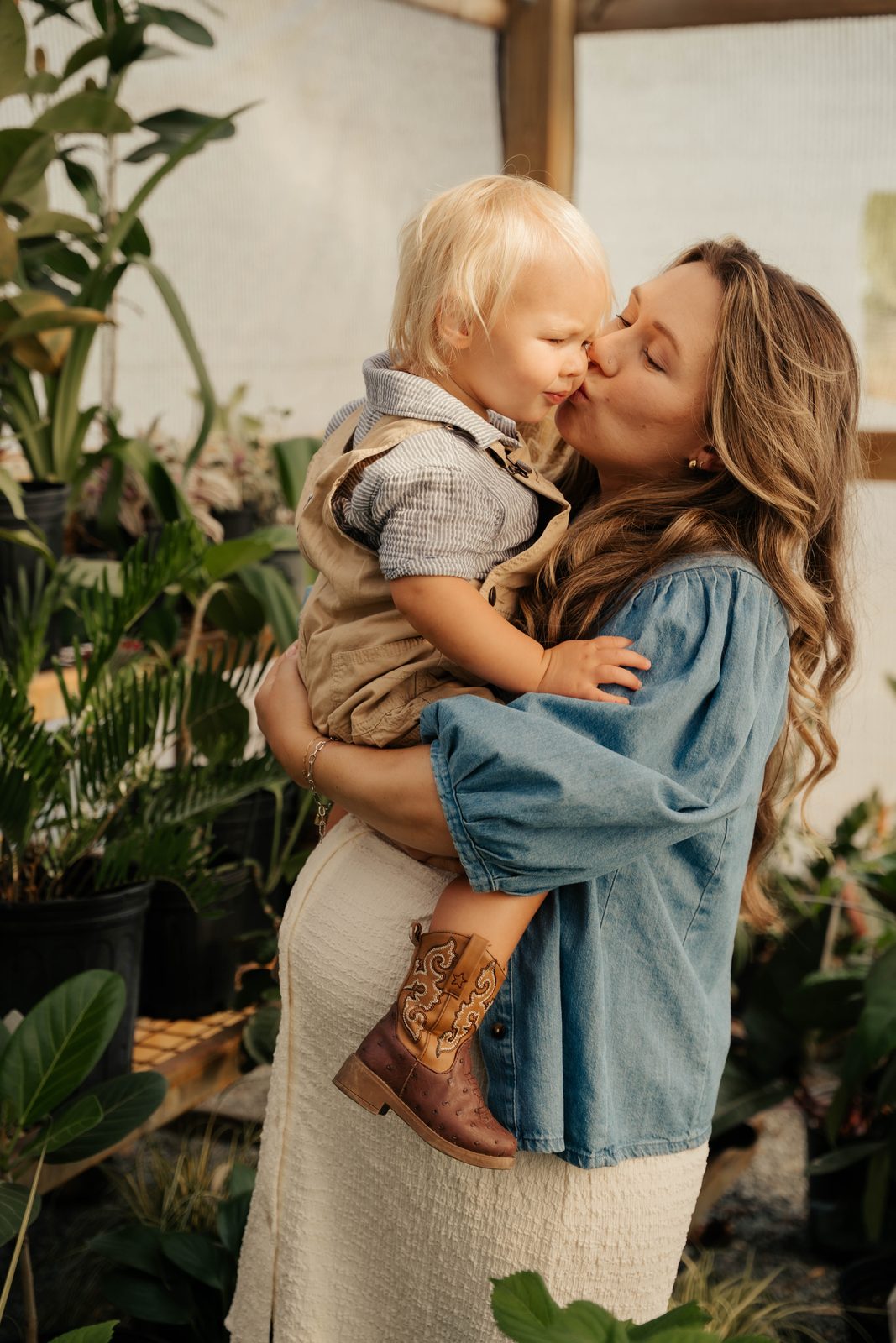 Mother kissing her toddler on the cheek while holding him in a greenhouse