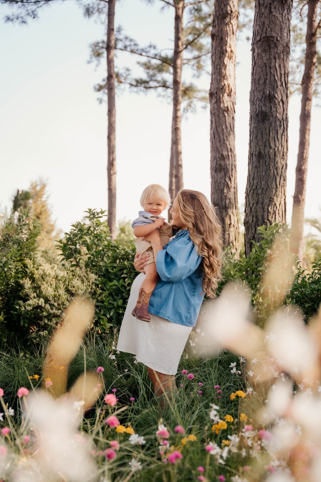 Mother in a denim shirt holding her toddler among tall pines and wildflowers
