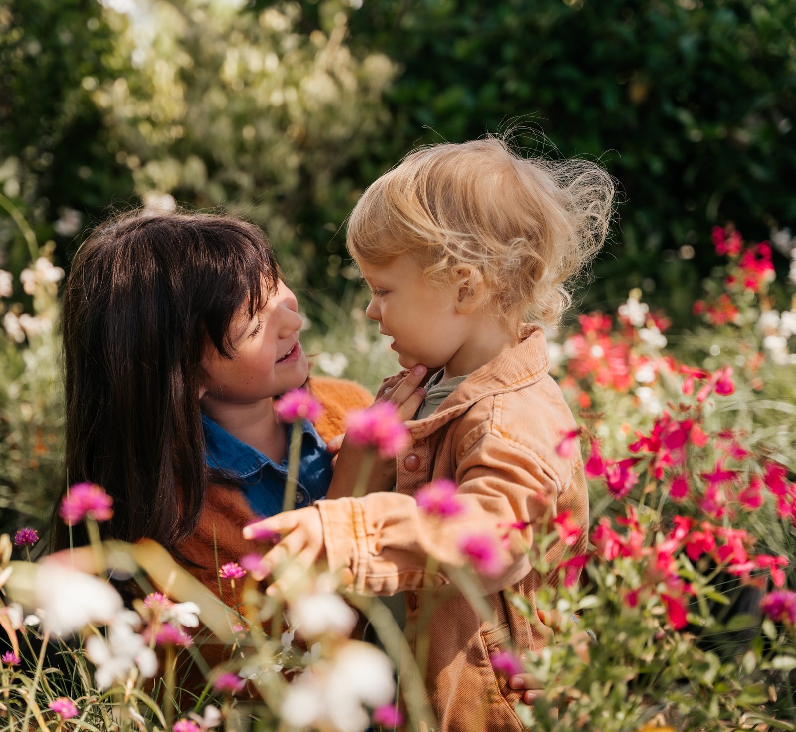 Older girl holding a blonde toddler face to face in a field of pink and white flowers