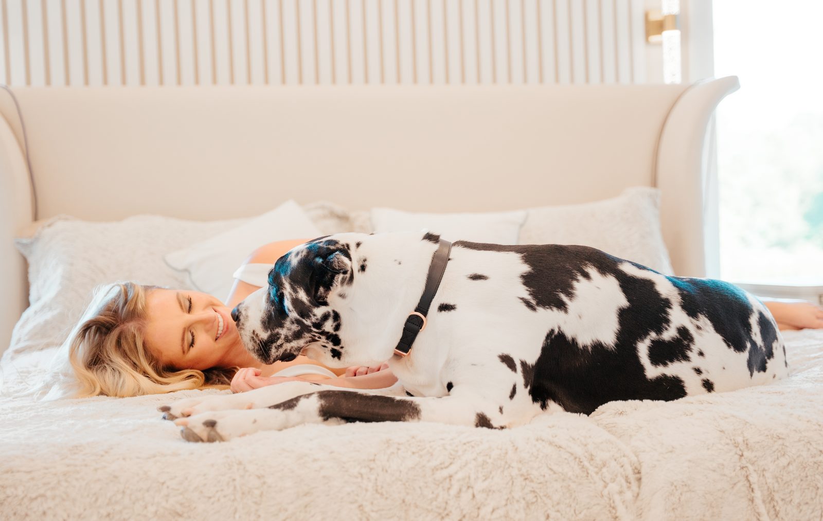 Woman lying on a white bed smiling as a Great Dane rests beside her