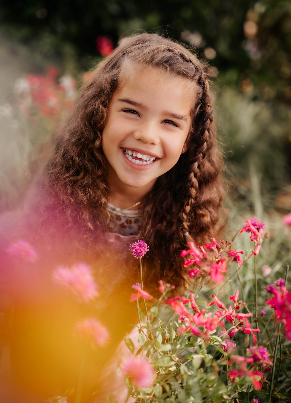 Young girl with curly hair and braids smiling widely among bright pink flowers