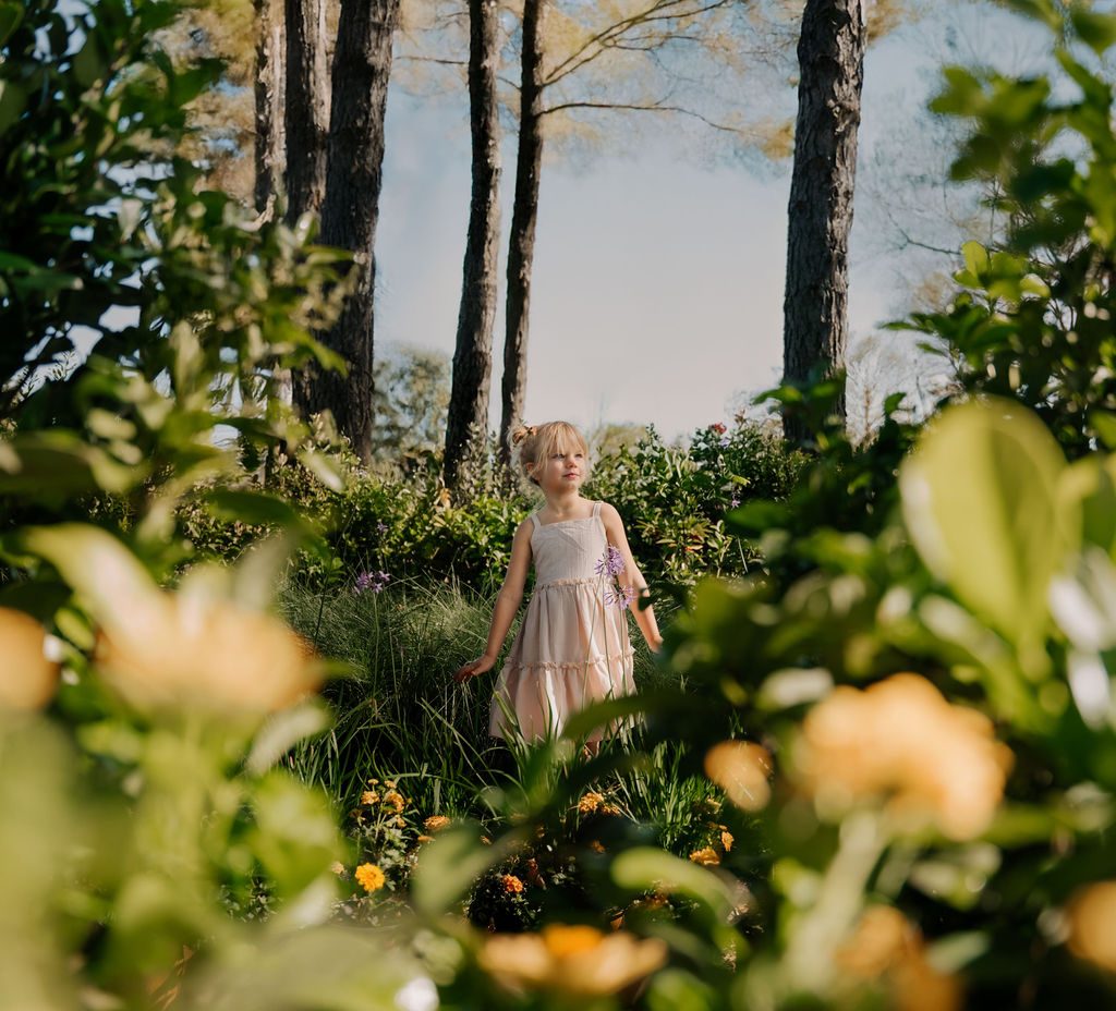 Girl in a cream dress standing among tall pines and yellow flowers