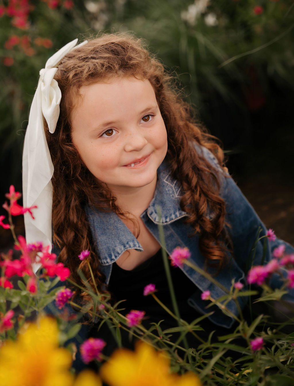 Girl with curly hair and a white bow smiling in a flower garden
