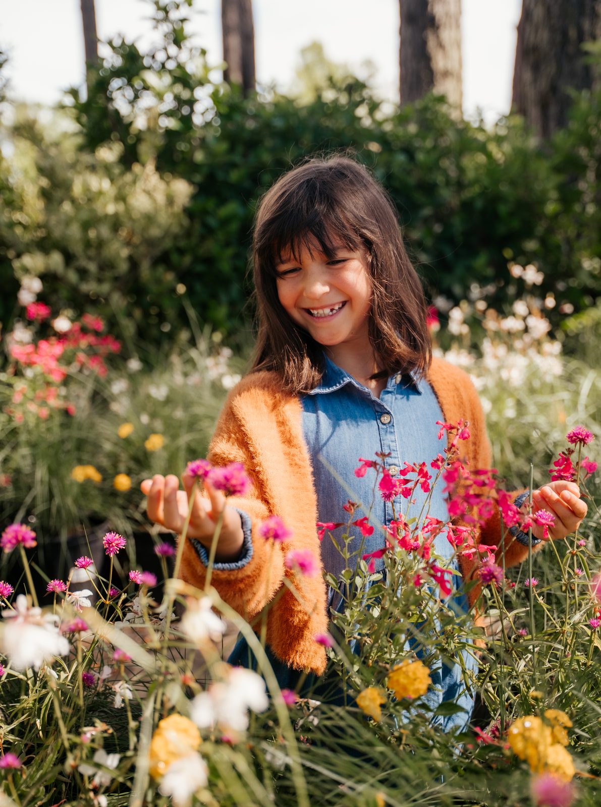 Girl in an orange cardigan and denim dress touching pink flowers with both hands
