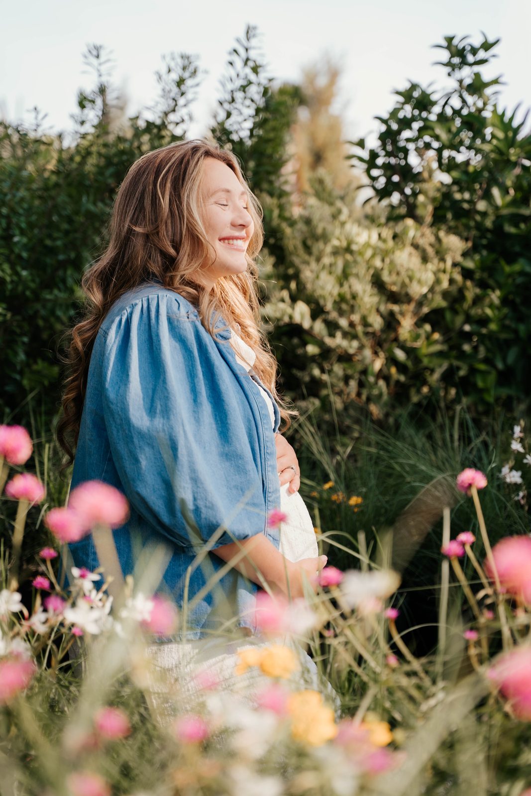 Expectant mother in a denim shirt resting her hands on her belly among pink wildflowers