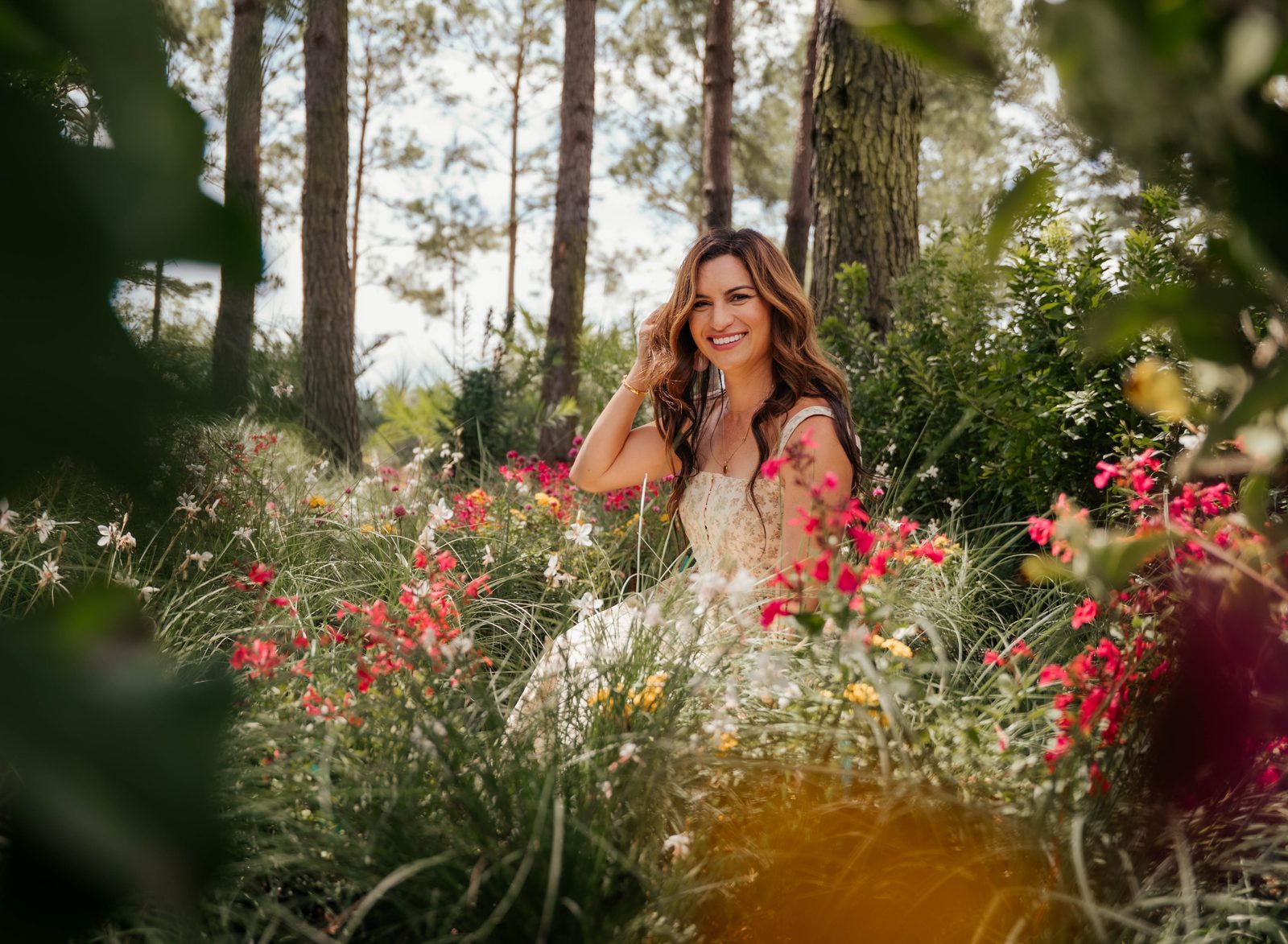 Woman in a cream floral dress smiling among wildflowers beneath tall pines
